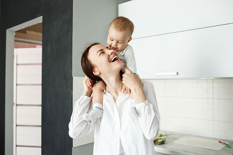 Mãe segurando seu bebe de cavalinho e sorrindo para ele para ilustrar o benefício maternidade