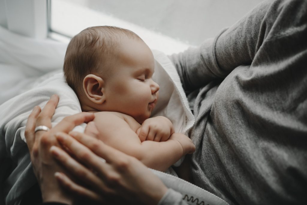 Bebê dormindo no colo de sua mãe e um close na câmera. Ambos estão de roupa cinza